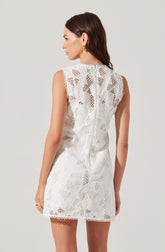 A woman is wearing a sleeveless white dress with intricate lace patterns. The dress features a high neckline with a scalloped edge and has a fitted silhouette that ends above the knees. The lace fabric displays geometric and floral cutouts, creating a delicate and elegant appearance.